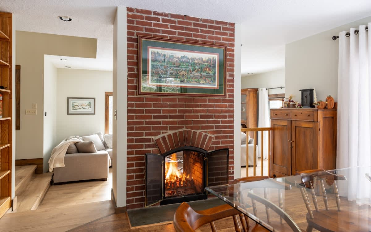 A close-up view of the brick fireplace in the dining room, featuring an arched opening with a roaring fire behind glass doors. A framed picture hangs above the mantel, and a glass-top dining table is visible in the foreground.