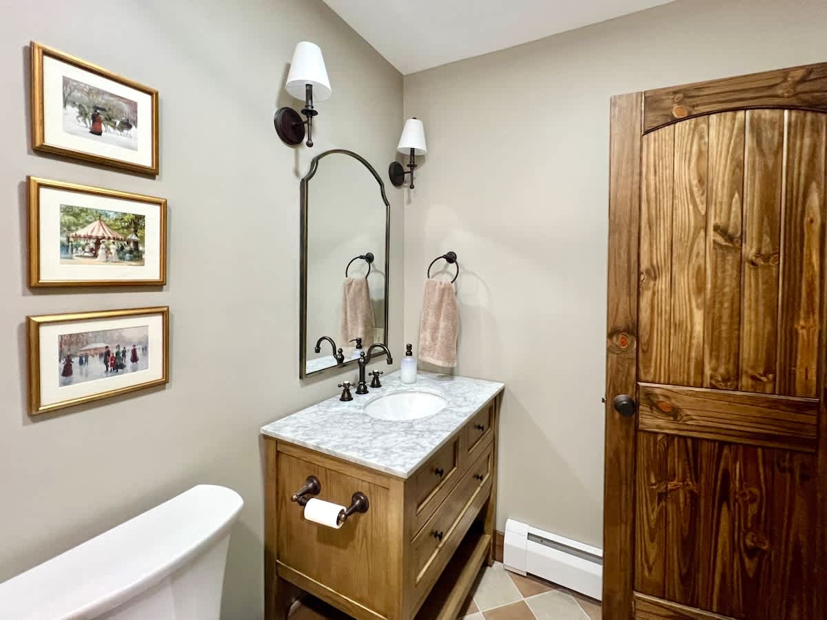 A half bathroom featuring a light wood vanity with a white marble countertop and sink, paired with a large arched mirror and sconce lighting. The room has light-colored walls, a toilet, and a rustic wooden door.