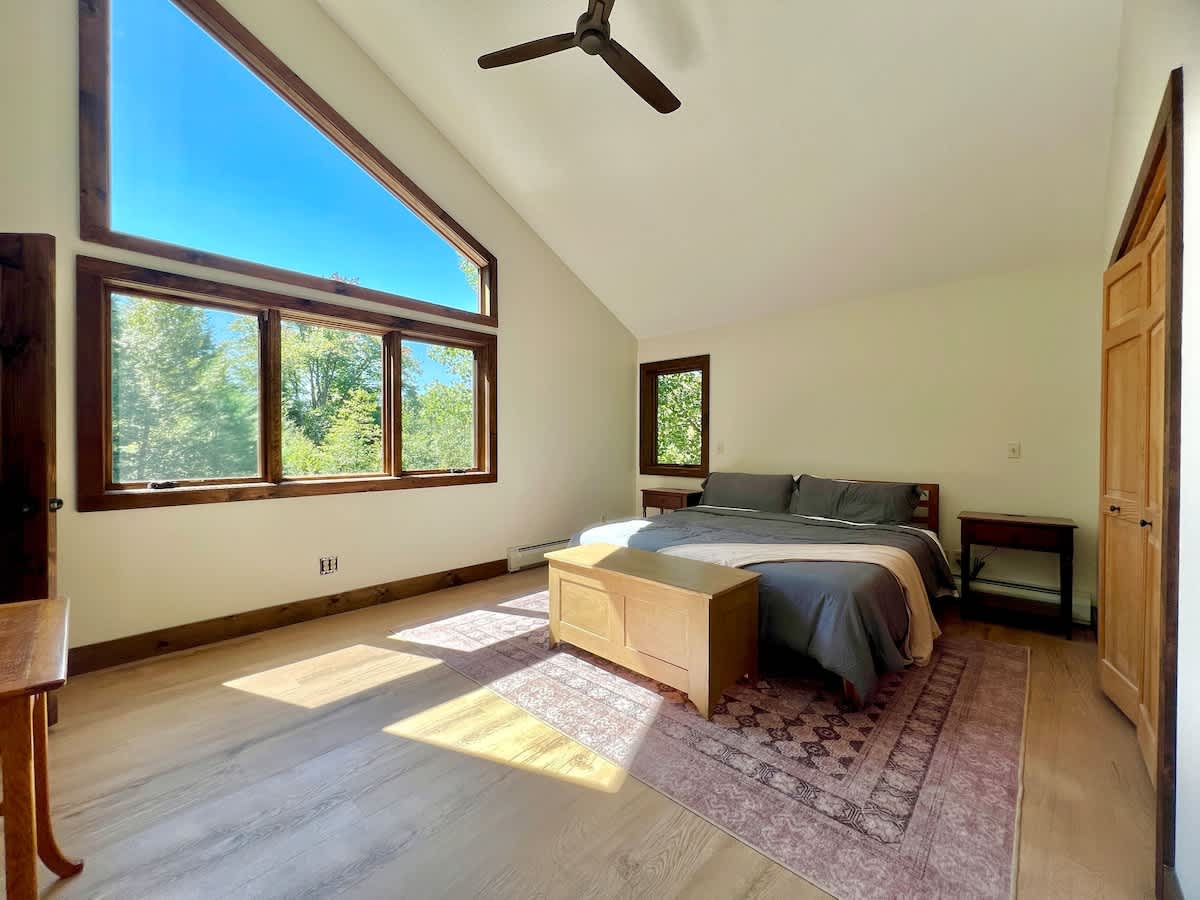 Bright and airy master bedroom with a high vaulted ceiling, large windows with wooden frames offering extensive forest views, light wood flooring, and a bed with grey bedding and a wooden storage chest.