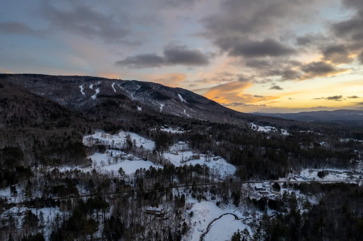Panoramic view of a snow-covered mountain landscape with ski slopes visible, under a dramatic sunset sky with orange and gray clouds. Dense forests cover the lower slopes and valleys.