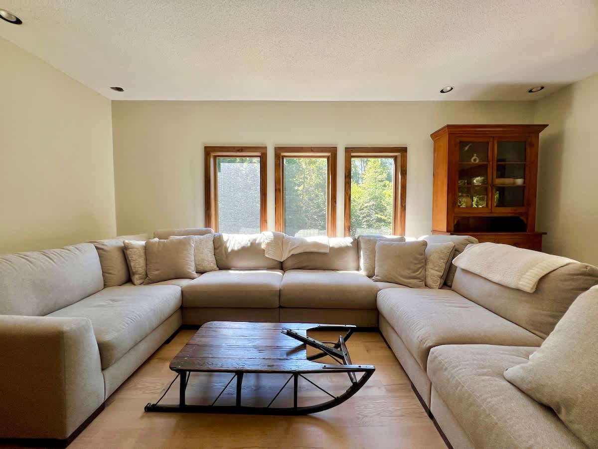 Spacious living room featuring a large, comfortable U-shaped sectional sofa in a light neutral fabric, centered around a unique wooden coffee table with a black sled-style base, illuminated by three large windows.