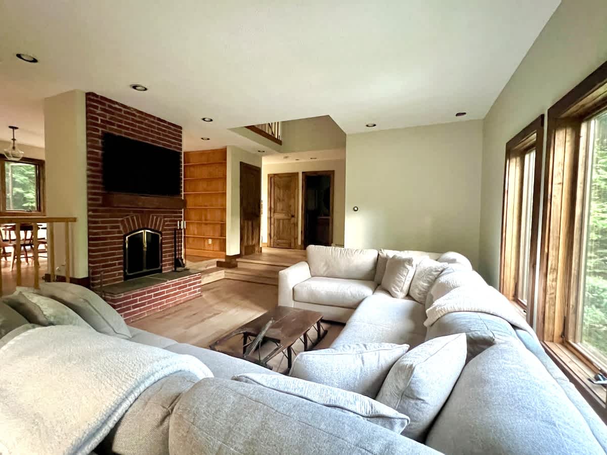 A spacious living room with a large L-shaped sectional sofa, hardwood floors, and a prominent brick fireplace with a TV mounted above. Large windows provide natural light, and a wooden built-in bookshelf is visible in the background.