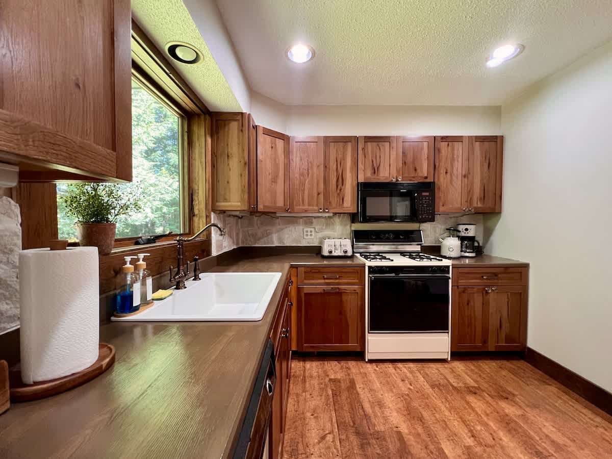 Well-appointed kitchen featuring warm wooden cabinets, dark countertops, a white farmhouse sink beneath a large window overlooking the forest, and essential appliances including a gas range and dishwasher.