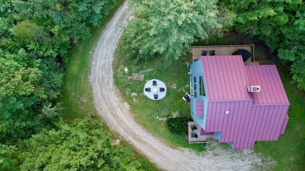 High aerial view of the blue house with its distinctive red metal roof, surrounded by dense green forest, revealing a curving gravel driveway and a circular outdoor fire pit area.
