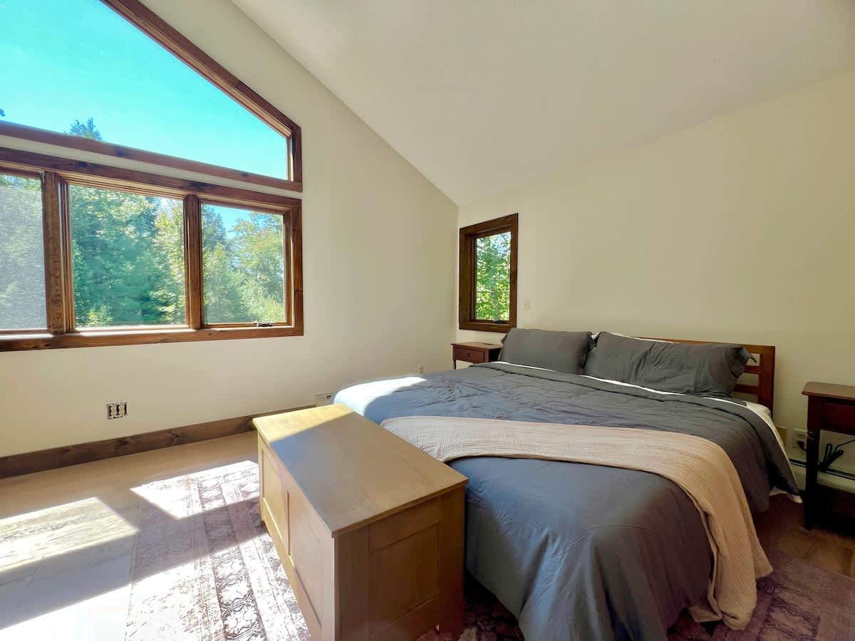 Spacious bedroom with a vaulted ceiling and expansive windows, including a large triangular window, showcasing lush forest views. Features a bed with grey bedding, a wooden trunk, and a patterned area rug.