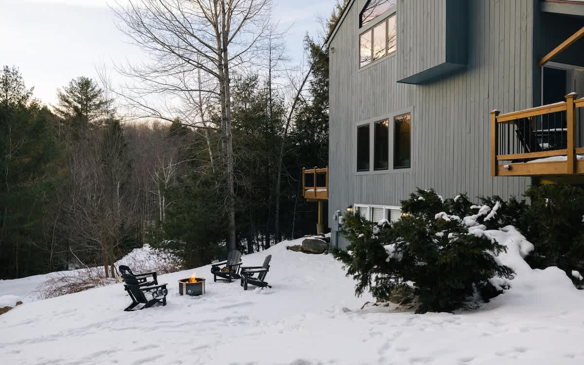 Wider view of the cabin's snowy backyard, showing the gray-blue exterior, a wooden deck, and a fire pit area with Adirondack chairs nestled among trees. The surrounding forest is visible under a bright sky.