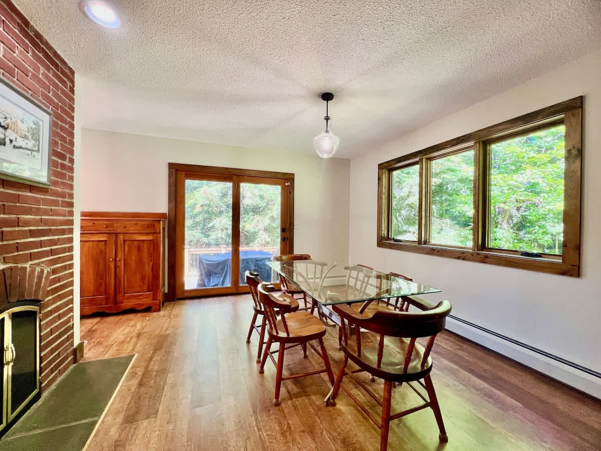 A bright dining room featuring a glass-top table with six wooden chairs, set on warm hardwood floors. A brick fireplace is visible on the left, and large windows and a sliding glass door offer views of the lush outdoors.