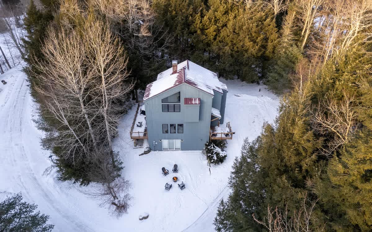 Aerial view of a gray-sided house with a red roof, surrounded by snow-covered evergreen and bare deciduous trees. A winding snowy path leads to the house, and a fire pit area is visible in the backyard.