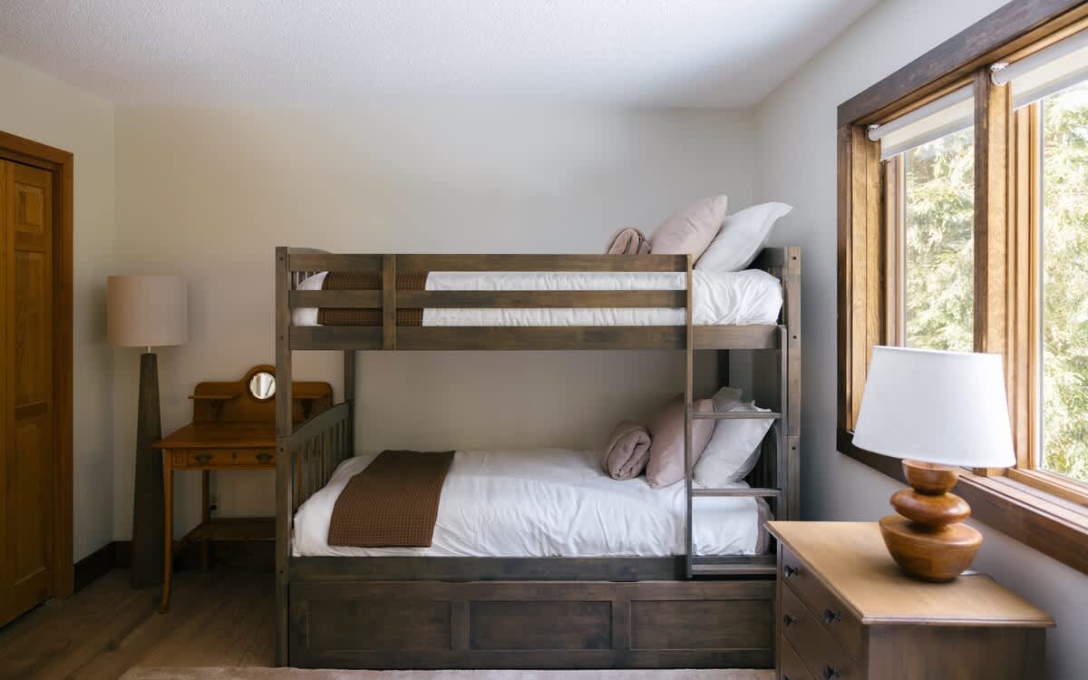 A closer view of the dark wood bunk bed with a trundle in bedroom-2, featuring white bedding and pink accent pillows. A small wooden desk and a floor lamp are visible to the left.