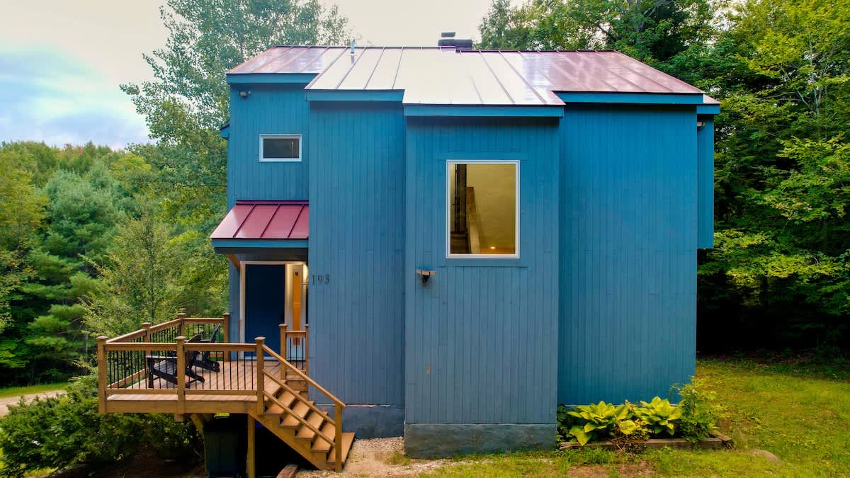 Striking blue multi-story house with a vibrant red metal roof, featuring a welcoming wooden deck with black railings and stairs leading to the main entrance, nestled among green trees.