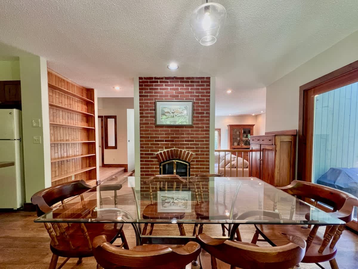 The dining area features a glass-top table with wooden chairs, positioned near a brick fireplace with an active fire. Built-in wooden shelves are visible on the left, and a sliding glass door leads to the outside deck.