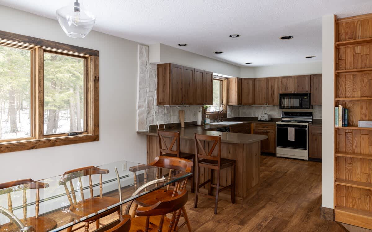 Open-concept kitchen and dining area featuring warm wood cabinetry, dark countertops, and a glass-top dining table with wooden chairs. Large windows offer views of a snowy, wooded landscape.
