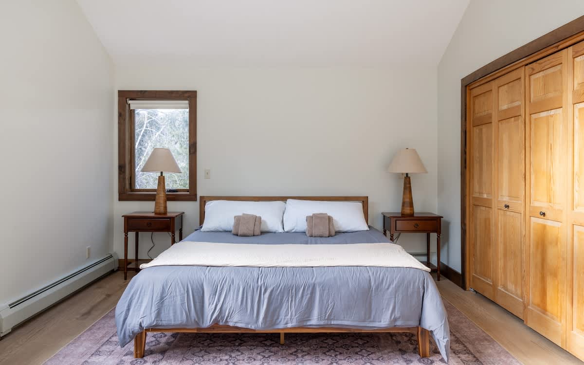 Bright bedroom with a queen-sized bed dressed in light blue linens and white pillows, flanked by two wooden nightstands with matching lamps. A large wooden closet and a window with rustic trim complete the space.