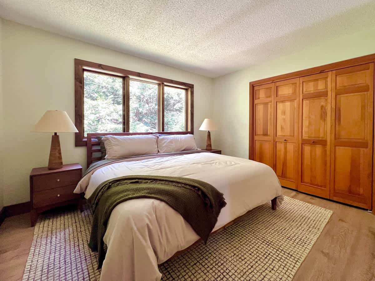 Cozy bedroom featuring a queen-sized bed with light bedding, two wooden nightstands with lamps, rectangular windows with wooden frames overlooking trees, and a large wooden closet.