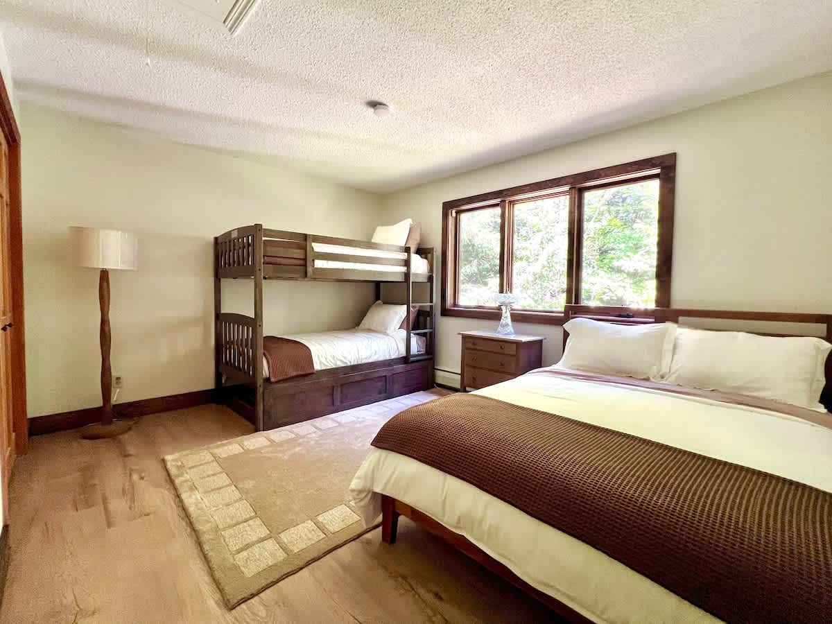 Spacious bedroom designed for multiple guests, featuring a queen bed with white bedding and a brown throw, and a sturdy wooden bunk bed. Windows with wooden frames offer natural light and forest views.