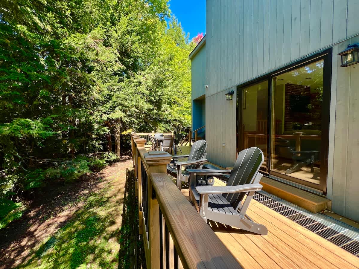 Inviting wooden deck furnished with two grey Adirondack chairs, offering a serene view of the surrounding lush green forest, accessible via a large sliding glass door from the house.