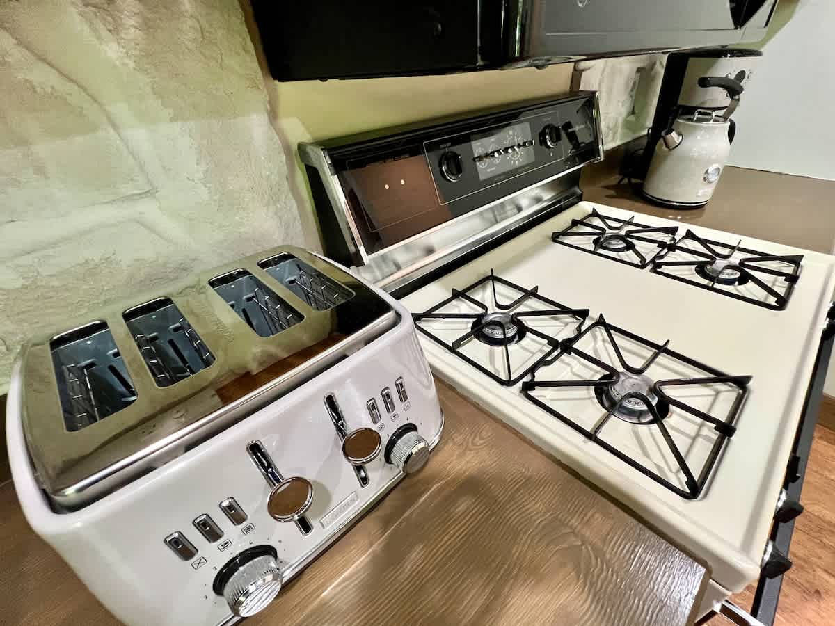 Close-up view of kitchen appliances, including a white gas range with black accents, a four-slice toaster, and a coffee maker, set against a wooden countertop and stone backsplash.