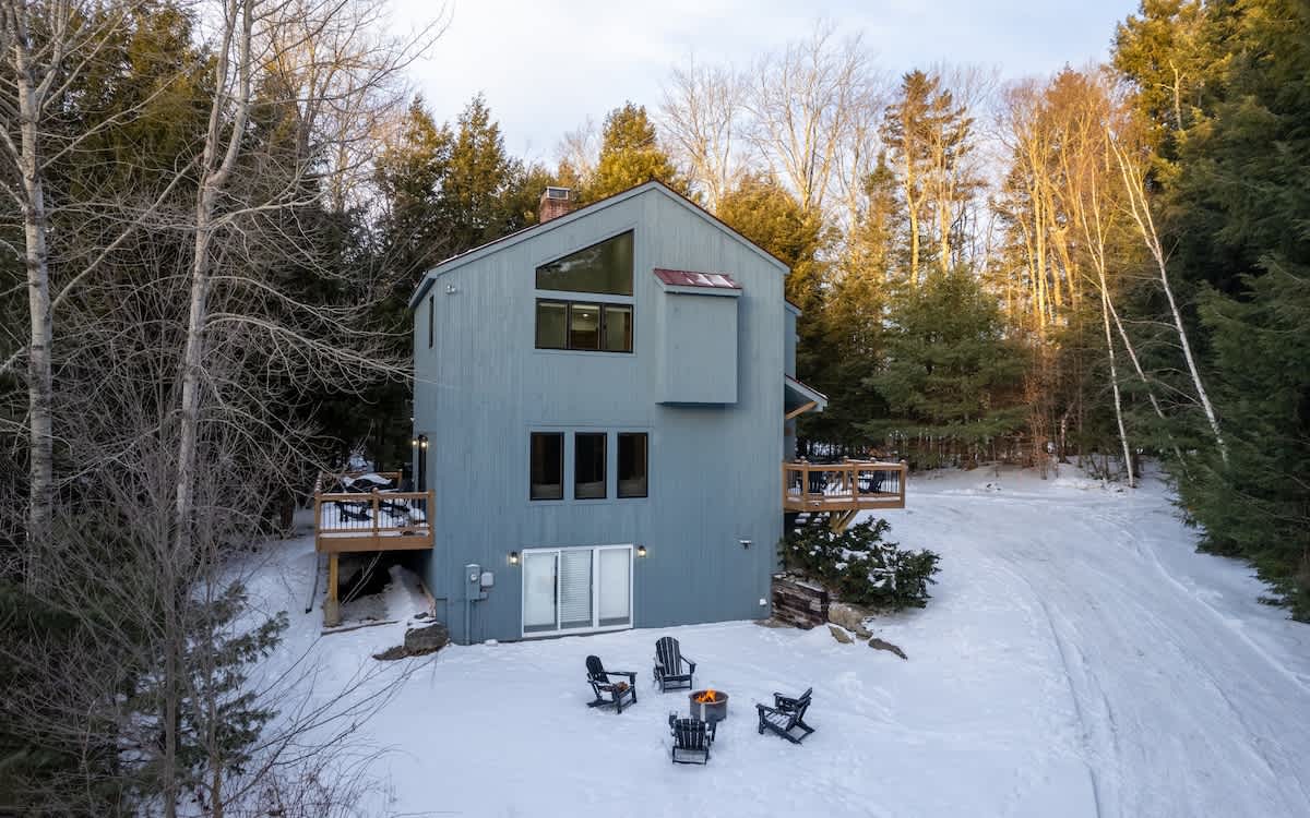 A modern gray-sided house with a prominent triangular window and two wooden decks, nestled among snow-dusted evergreen and bare deciduous trees. A fire pit with four Adirondack chairs sits on the snowy ground.