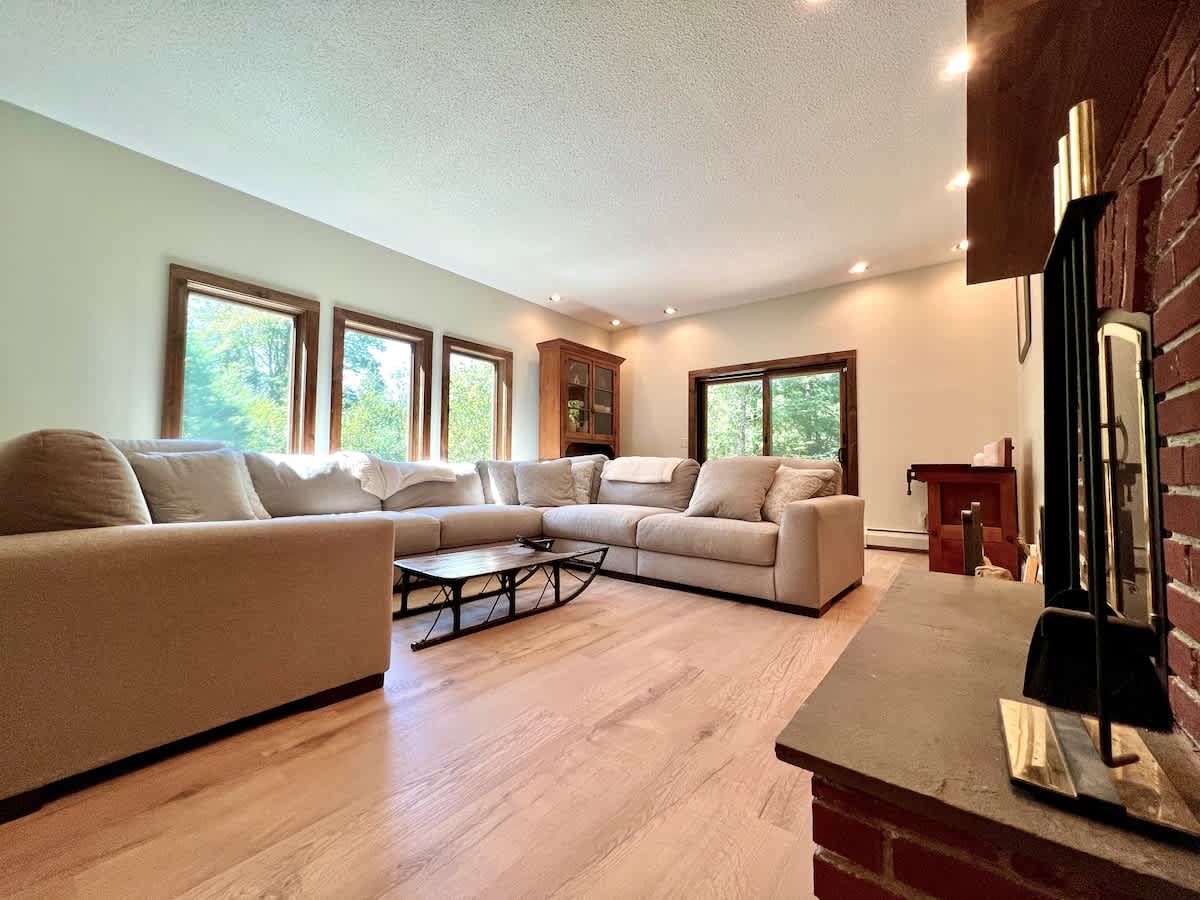 A spacious living room with a large L-shaped sectional sofa in a light neutral color, positioned on hardwood floors. A prominent brick fireplace with a mounted television serves as a focal point, complemented by large windows with wooden trim offering outdoor views.