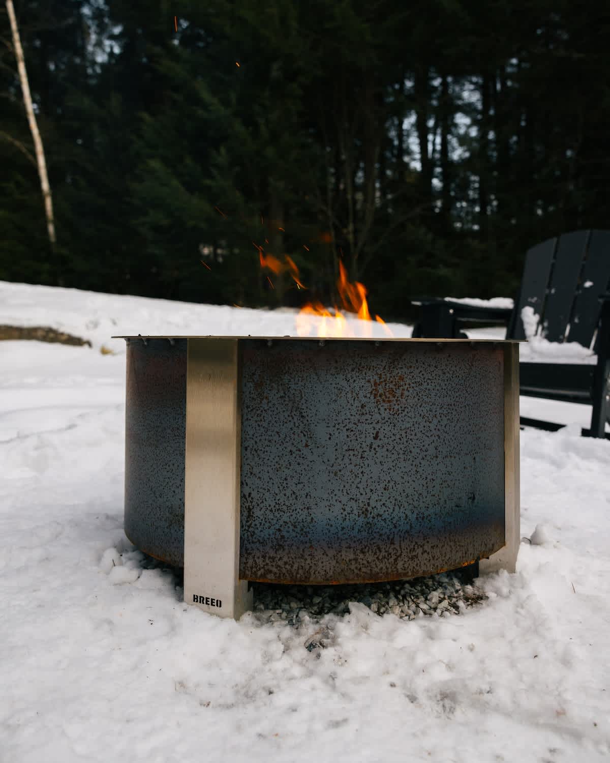 Close-up of a modern, cylindrical metal fire pit with flames burning, surrounded by snow and gravel. The brand "BREEO" is visible on the side of the fire pit.