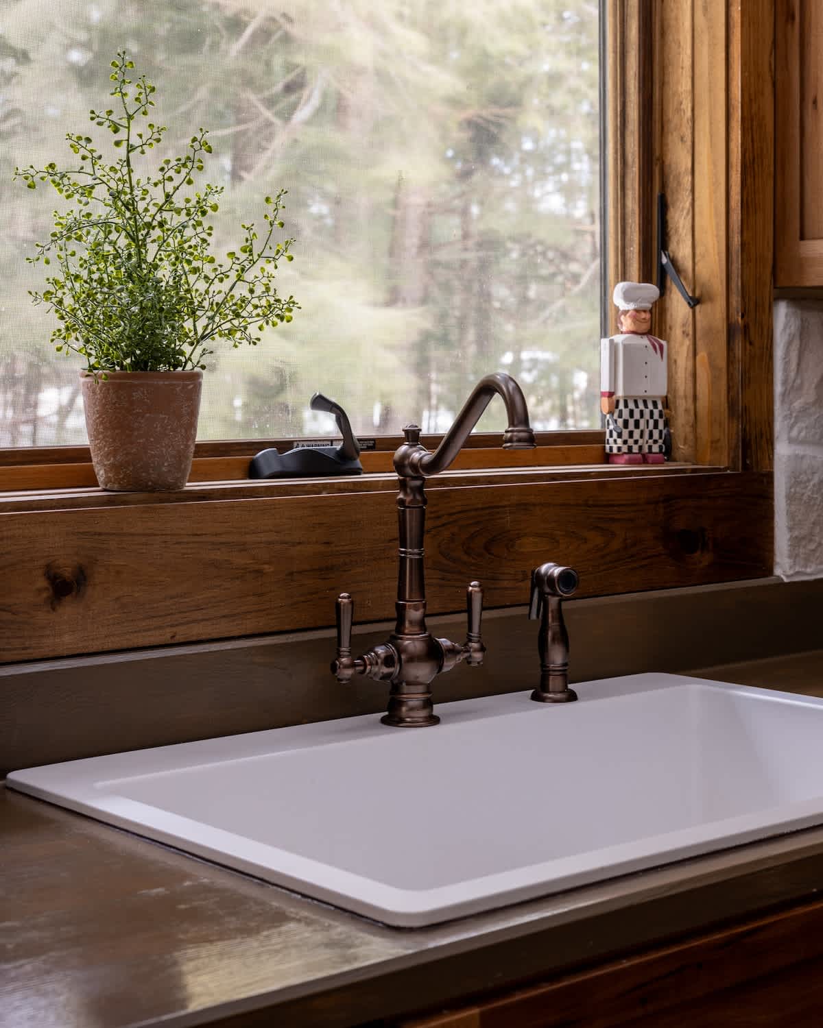 Close-up of a white farmhouse sink with a bronze faucet and sprayer, set against a dark countertop and rustic wood window trim. A small potted plant and a chef figurine adorn the windowsill.