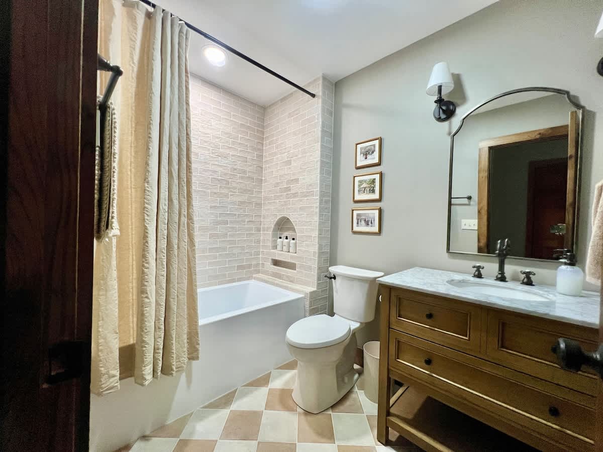A full bathroom featuring a white bathtub with light subway tile surround and a neutral shower curtain. A light wood vanity with a marble top and sink is on the right, complemented by an arched mirror and sconce lighting, all set on a checkered tile floor.