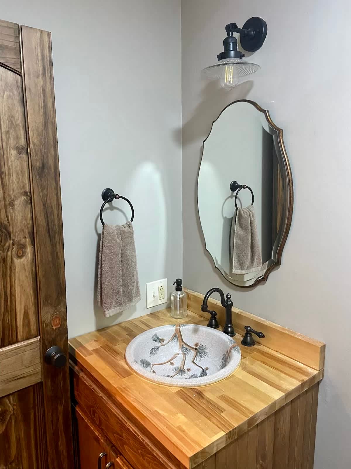 A half bathroom featuring a unique vessel sink with a nature-inspired design set on a wooden countertop and vanity. An oval mirror with a dark frame and a single sconce light are mounted above, next to a rustic wooden door.