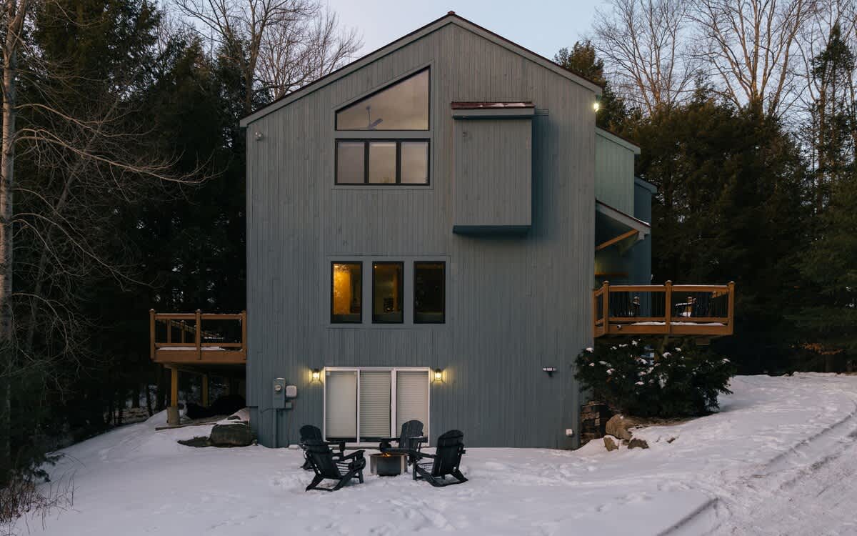 Modern gray-sided house with multiple windows and two wooden decks, set against dark evergreen trees. A fire pit with four Adirondack chairs sits on the snow-covered ground in the foreground under a clear sky.