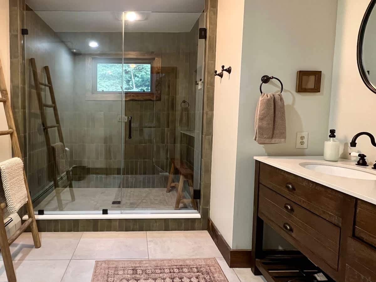 Modern bathroom with a large glass-enclosed shower featuring light brown subway tiles, a wooden ladder for towels, and a dark wood vanity with a white sink and black fixtures.