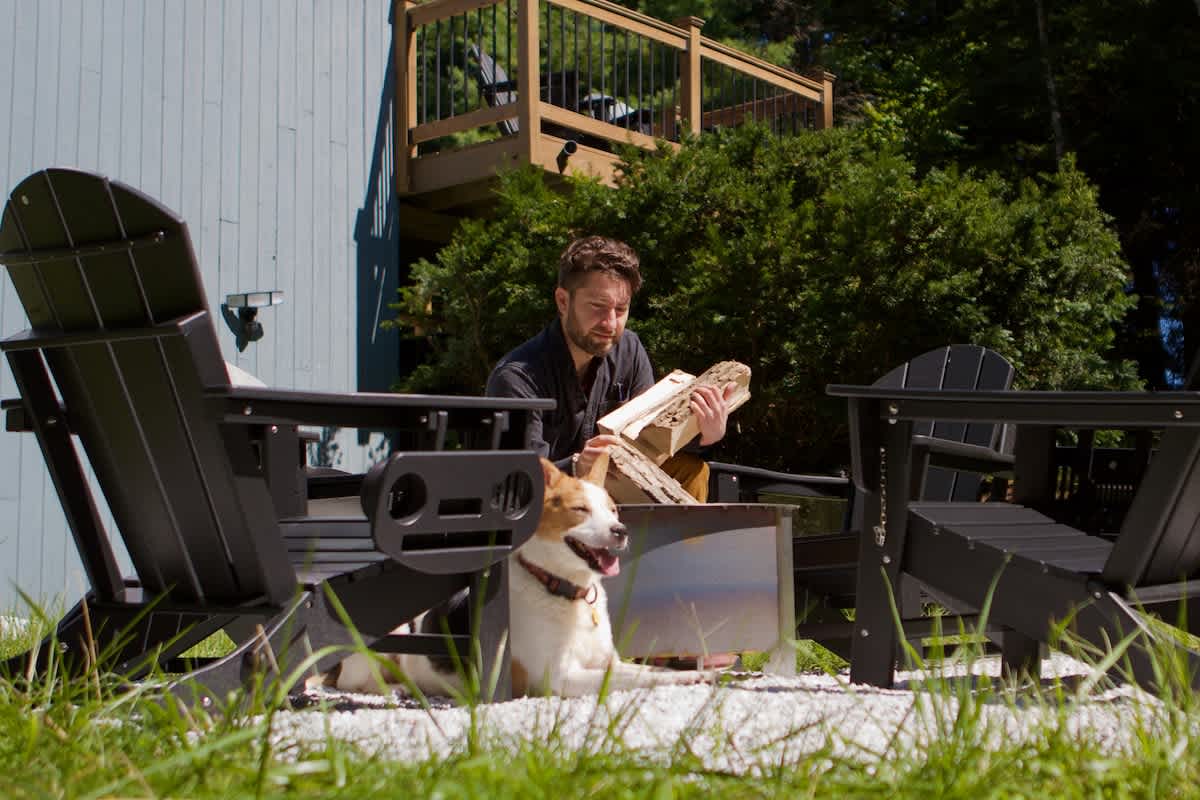 A man adds logs to a stainless steel fire pit, surrounded by black Adirondack chairs on a white gravel patch, with a happy dog resting nearby and a house deck in the background.