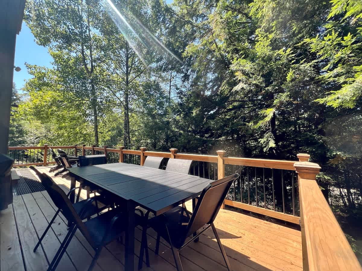 Spacious outdoor wooden deck surrounded by lush green trees, featuring a long black dining table with six chairs. The deck has wooden railings with black balusters, perfect for outdoor dining.