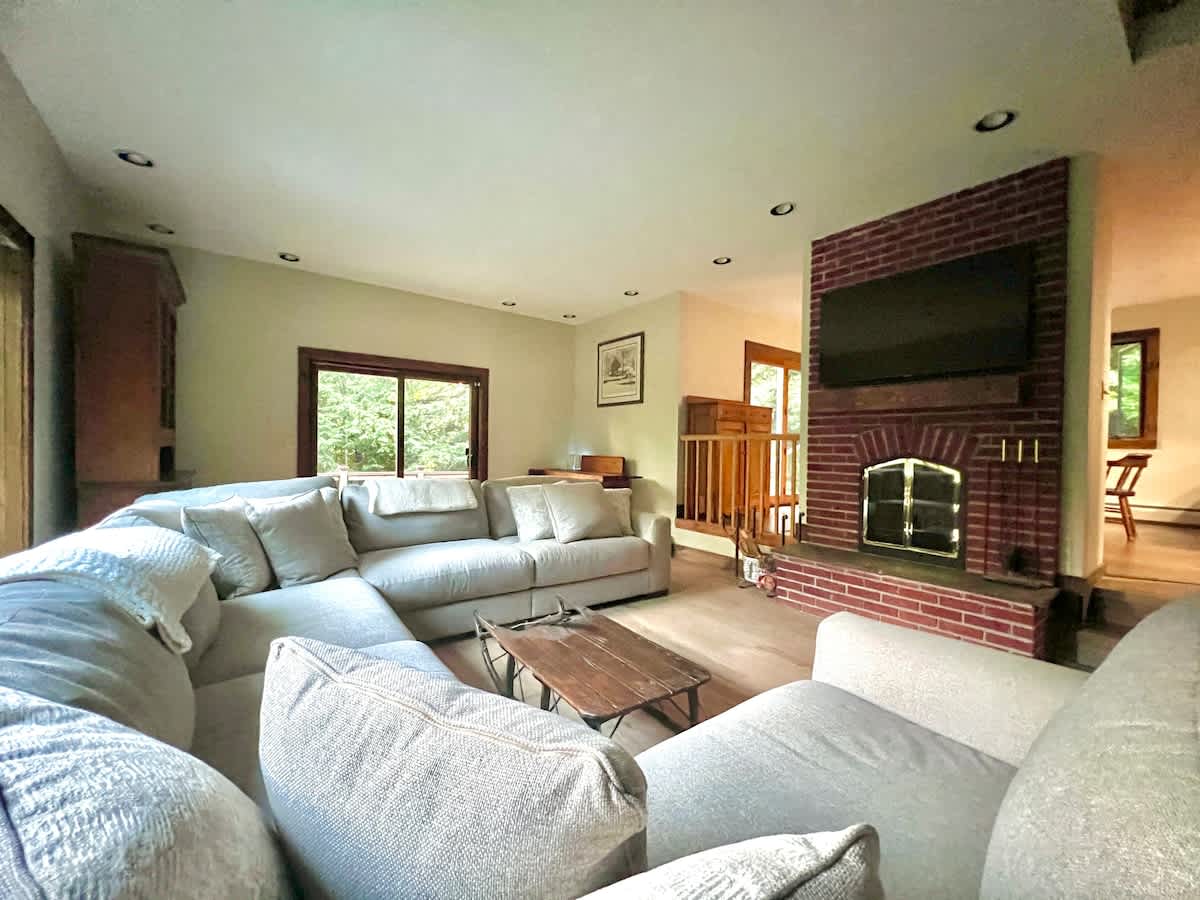 A cozy living room featuring a large L-shaped sectional sofa in a light color, centered around a brick fireplace with a television mounted above. Hardwood floors extend throughout the space, and natural light streams in from large windows.