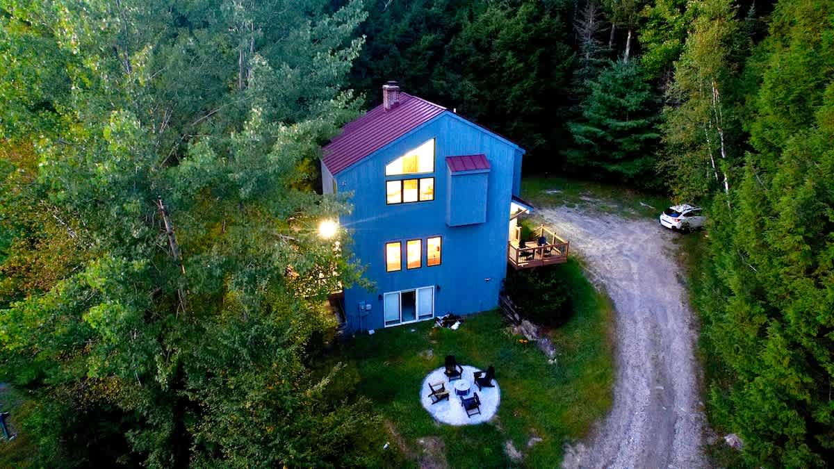 Expansive aerial view of the blue house with a red metal roof, nestled within a dense green forest, showcasing a long gravel driveway, an outdoor fire pit area, and warm interior lights.