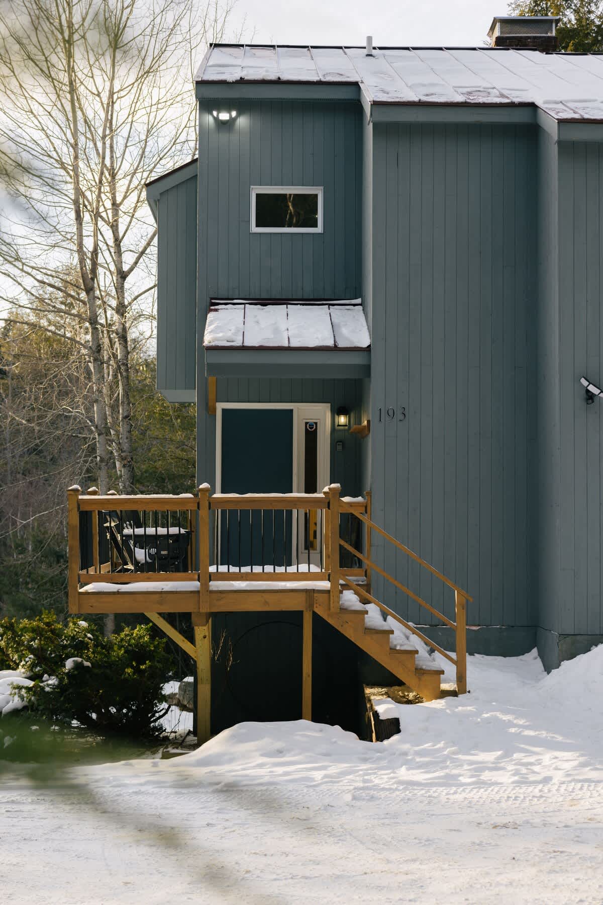Front exterior of a gray-blue cabin with a wooden deck and stairs leading to the entrance, partially covered in snow. The house number "193" is visible next to the door.