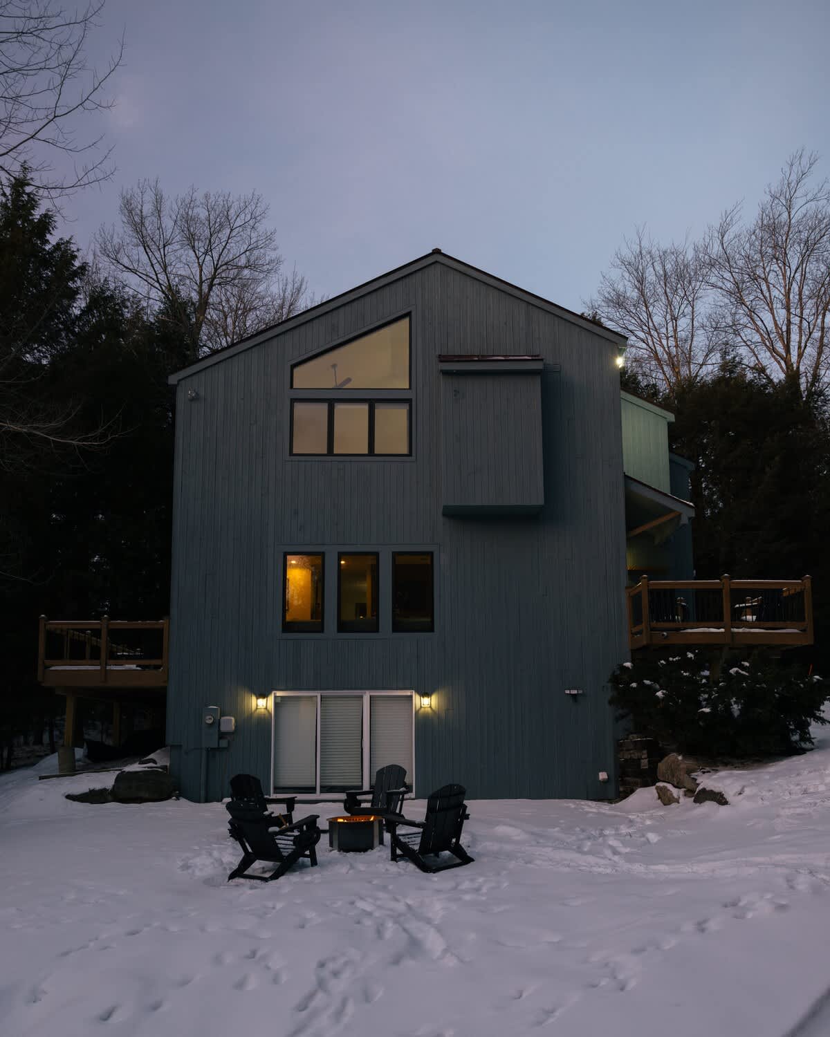 The back of a modern gray-sided house at dusk, with interior lights glowing through windows and exterior lights illuminating a fire pit area with four Adirondack chairs on snow. Dark trees frame the house.