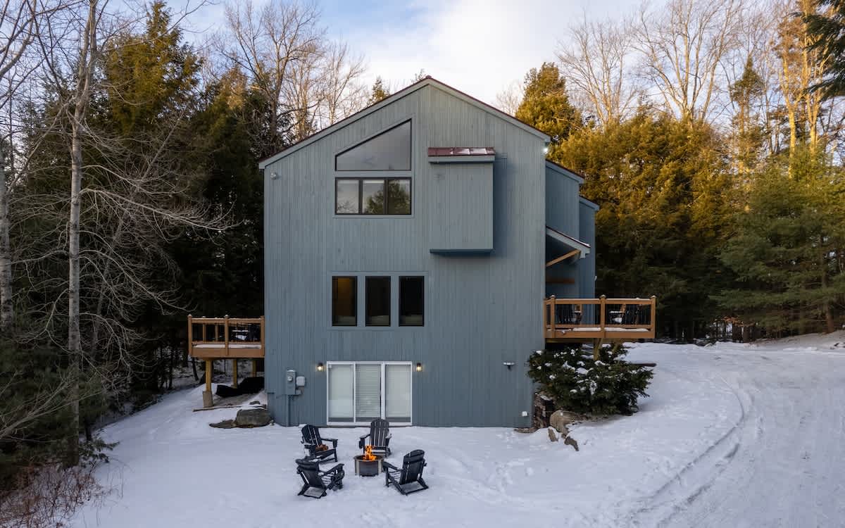 The back of a tall, modern gray-sided house featuring multiple windows and two wooden decks, surrounded by snow-covered trees. A fire pit with four Adirondack chairs is arranged on the snowy ground.
