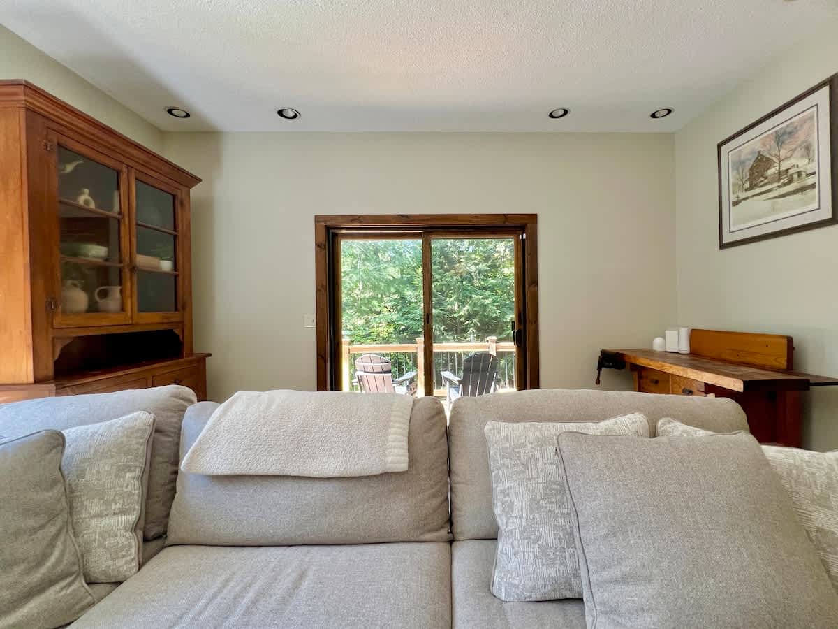 Comfortable living room with a large light-colored sectional sofa, a sliding glass door leading to an outdoor deck with forest views, recessed lighting, and rustic wooden furniture including a hutch and desk.