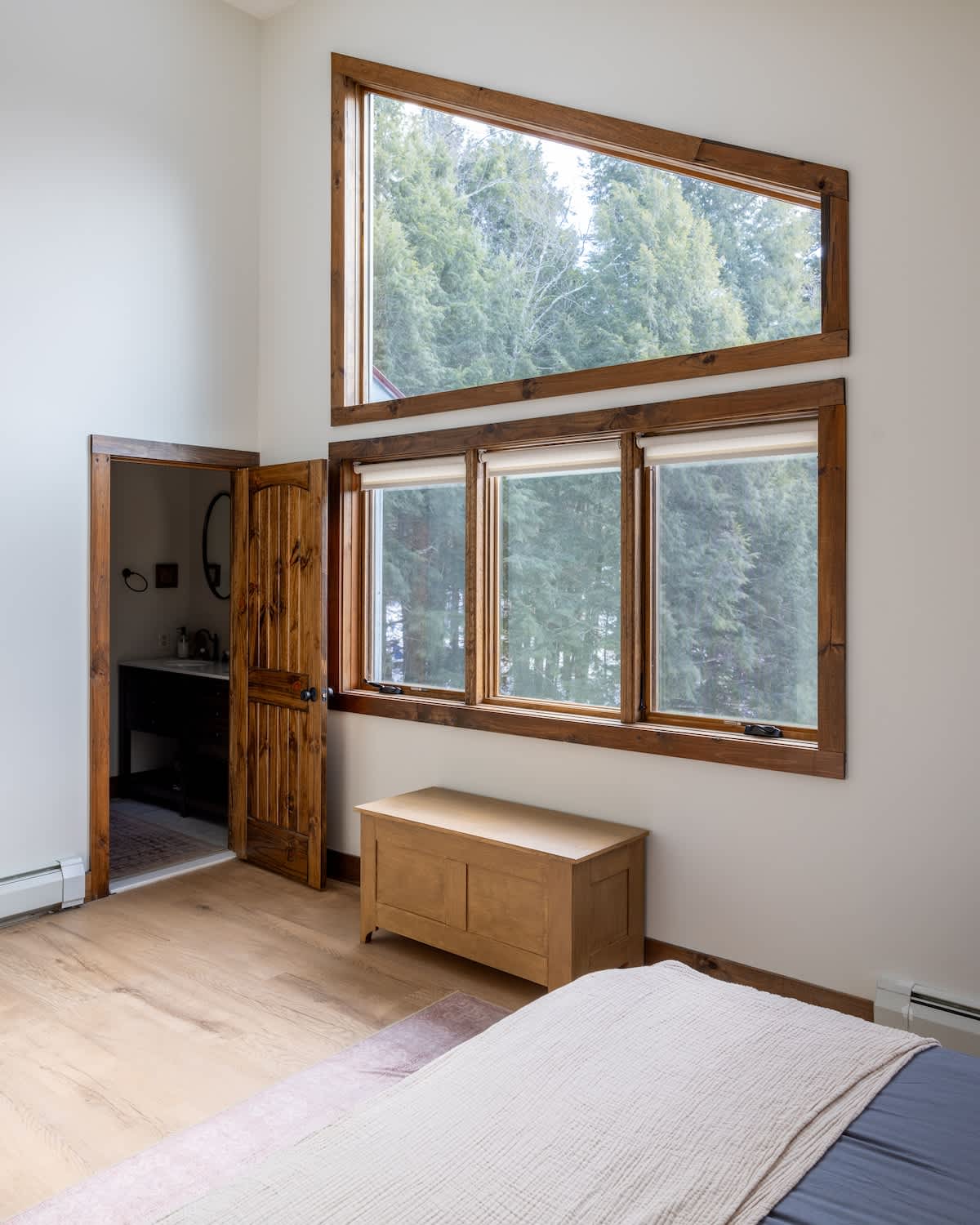 Another view of bedroom-1, showcasing a unique triangular window above a set of three rectangular windows, all with rustic wood frames. A wooden chest sits below the windows, and a bathroom door is visible to the left.