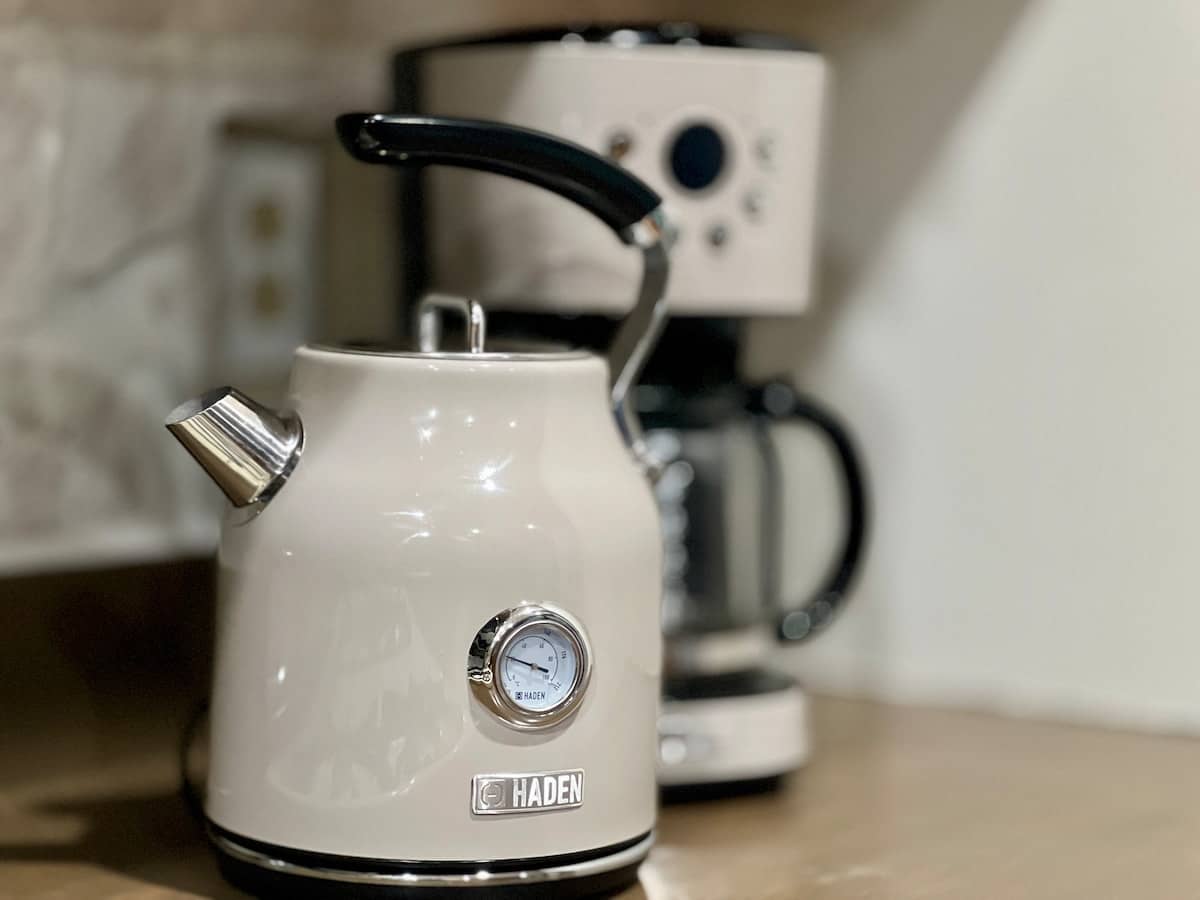 Close-up of a cream-colored retro electric kettle with a temperature gauge, positioned on a kitchen counter. A matching coffee maker is visible in the blurred background, suggesting a well-equipped kitchen.