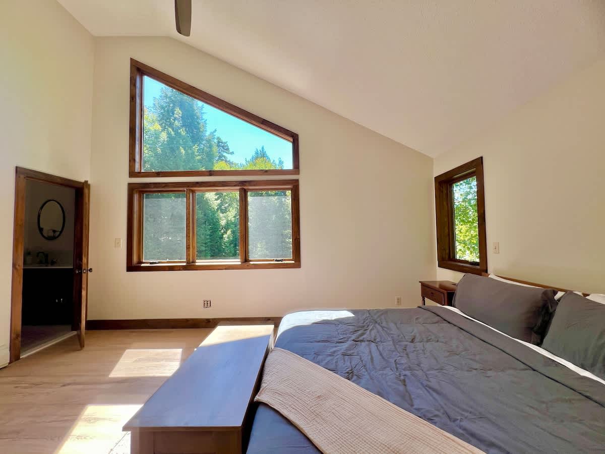 Bright master bedroom with a vaulted ceiling, large triangular and rectangular windows framed in wood offering forest views, light wood floors, and a bed with grey bedding and a wooden bench.