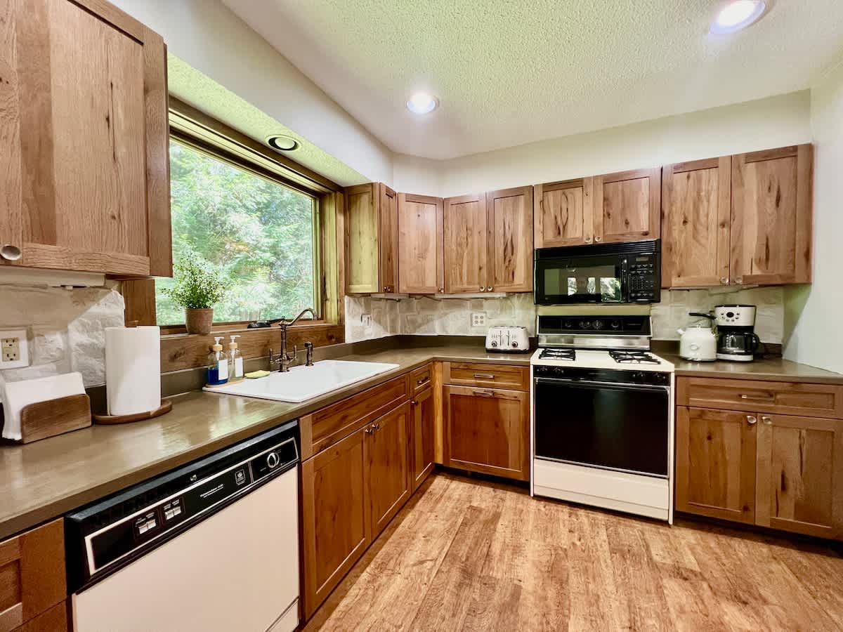 Fully equipped kitchen with natural wood cabinetry, dark countertops, a large white farmhouse sink under a window with forest views, a white gas range, and integrated dishwasher.