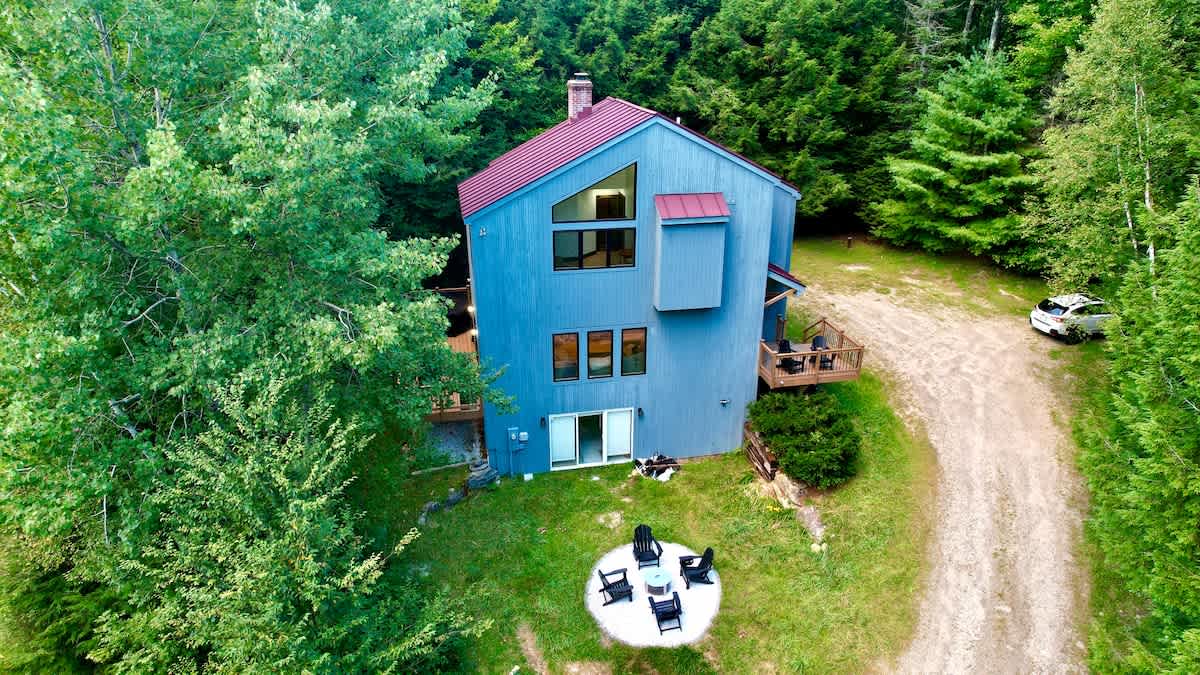 Overhead aerial shot of the blue house with a red metal roof, surrounded by a vibrant green forest, highlighting a winding gravel driveway and a distinct circular fire pit area with seating.
