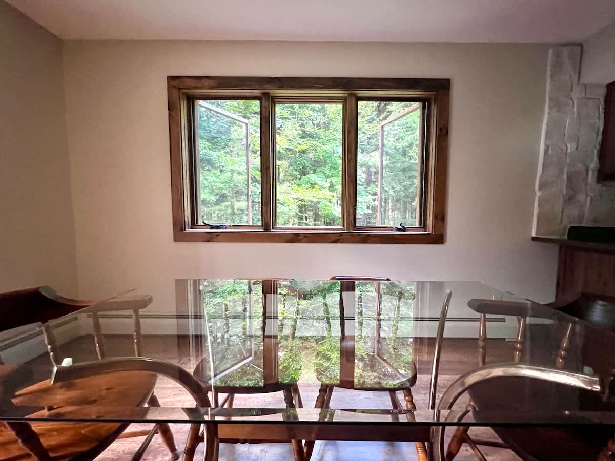 Dining area with a clear glass-top table and six wooden chairs, set against a light-colored wall. A large window with wooden trim offers a view of the surrounding forest, bringing nature indoors.