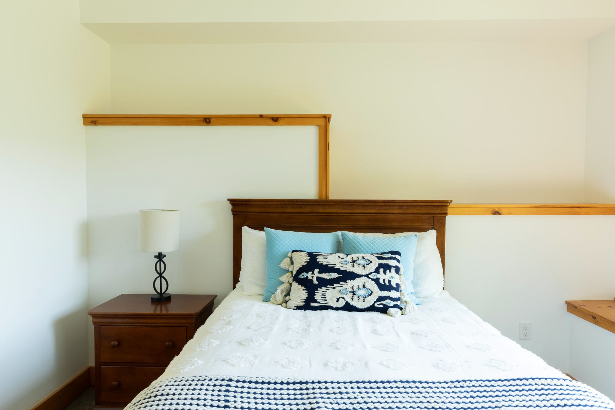 A close-up view of a dark wood bed dressed in a white textured comforter, a blue knitted throw, and a mix of blue and patterned decorative pillows, with a wooden nightstand and lamp beside it.