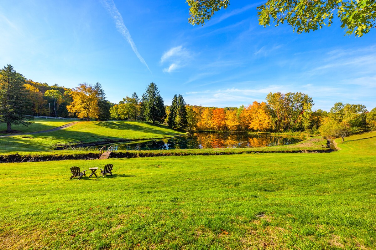 A picturesque landscape featuring a vibrant green lawn leading to a tranquil pond, surrounded by trees displaying brilliant autumn foliage under a clear blue sky. Two Adirondack chairs are visible by the pond.