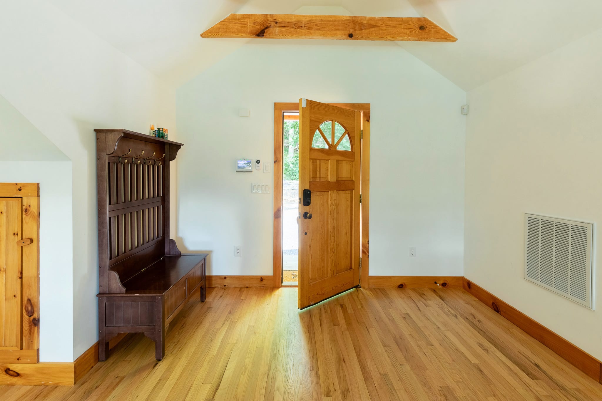 A bright and airy foyer with light hardwood floors, white walls, and a rustic wooden bench, featuring an open wooden door that leads to the outdoors.