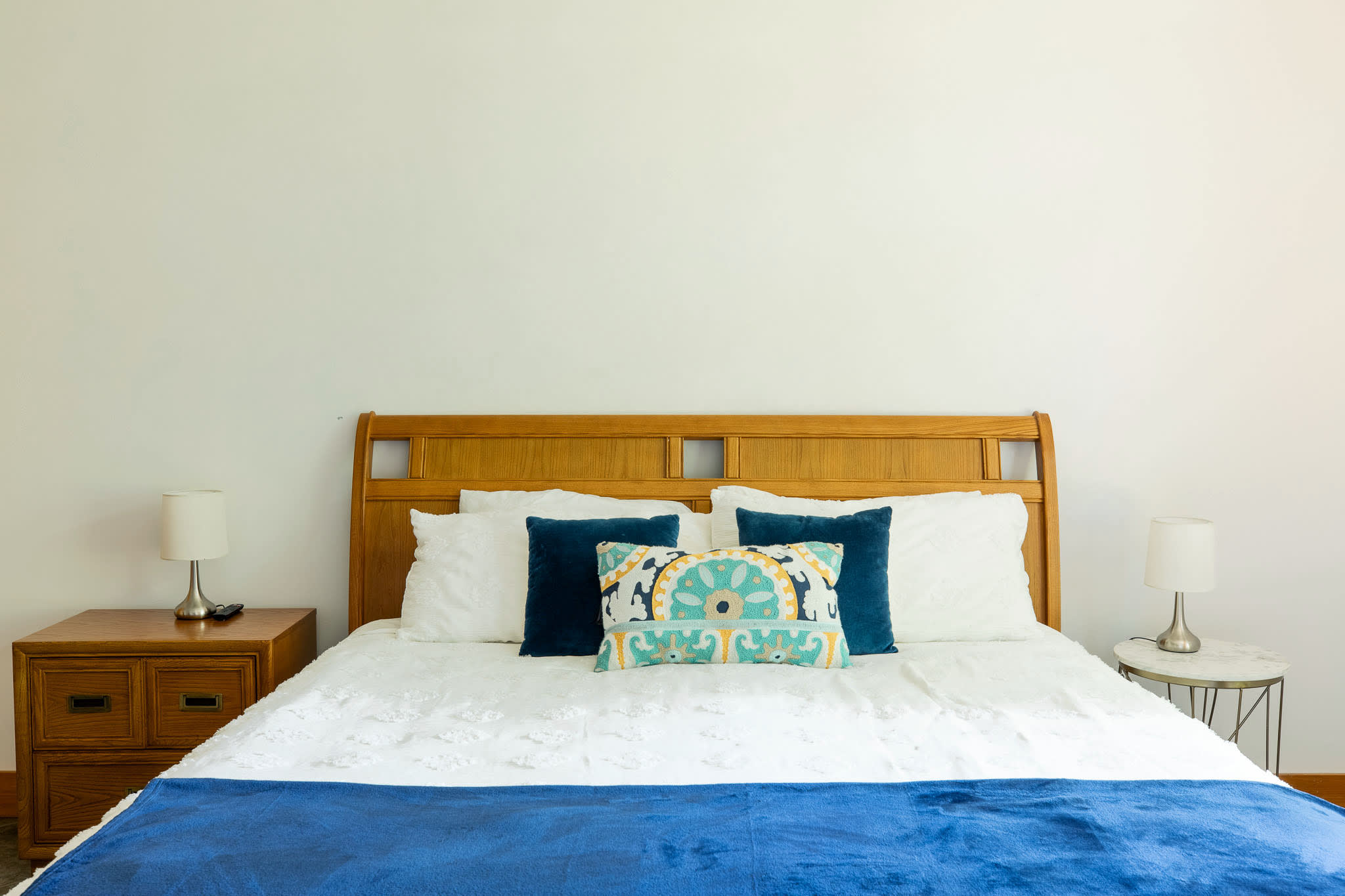 A close-up of a wooden queen-sized bed with a white comforter, a blue throw, and a mix of blue and patterned pillows, flanked by two wooden nightstands with lamps.