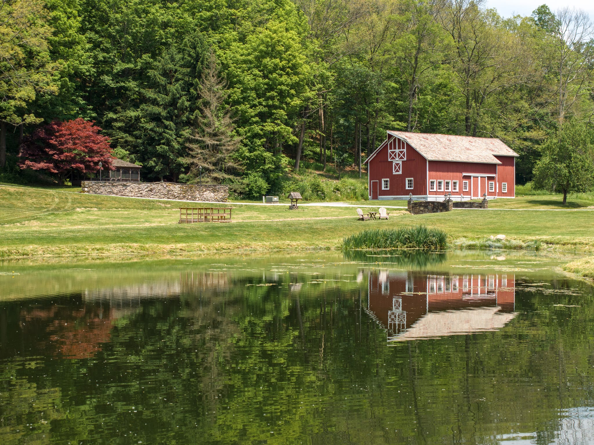 A picturesque red barn with white trim reflected in a calm pond, surrounded by expansive green lawns and dense forest, showcasing a rustic rural setting.