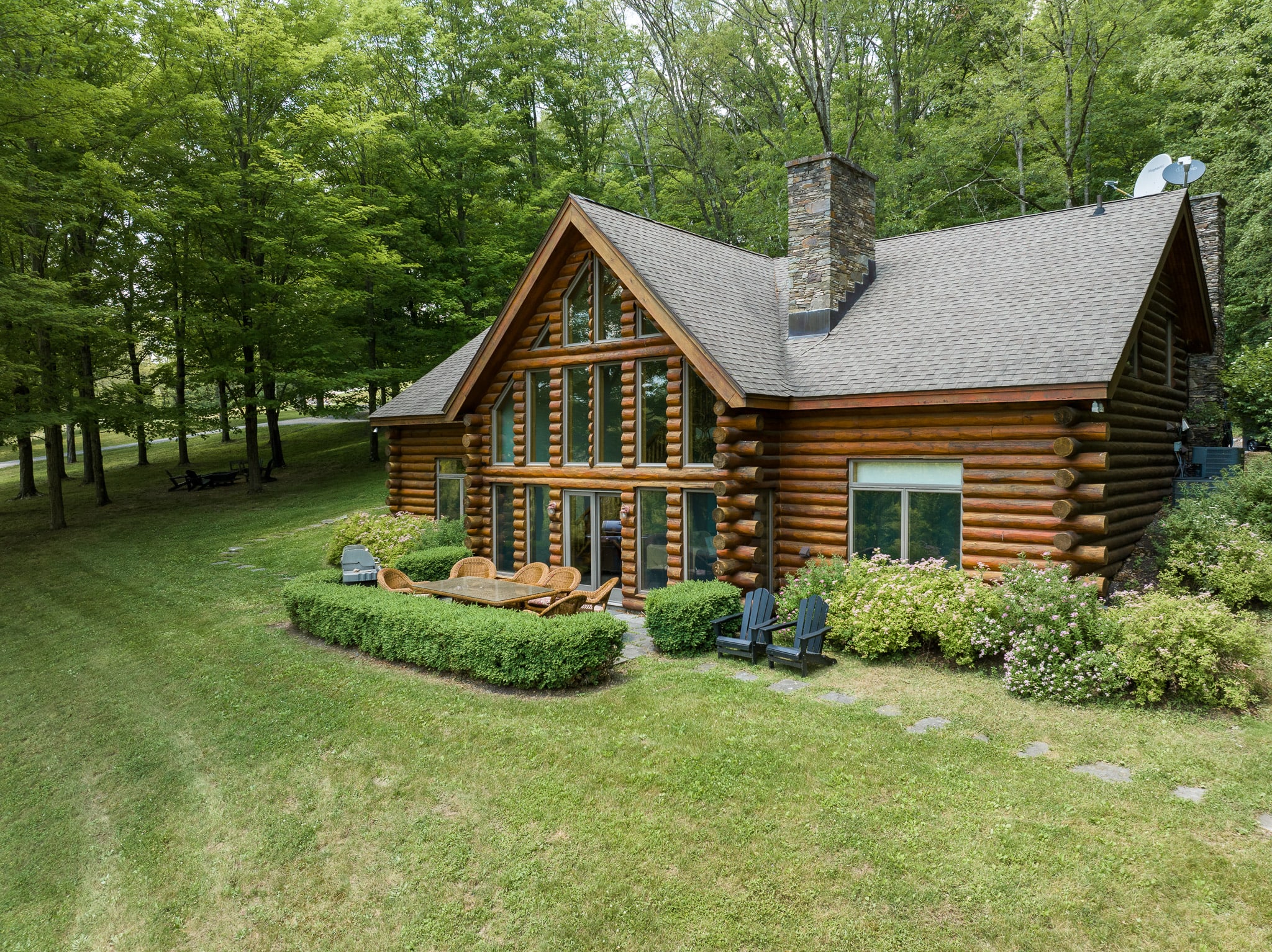 An aerial view of a rustic log cabin nestled among lush green trees, featuring a prominent stone chimney and a patio with outdoor seating and a BBQ grill.