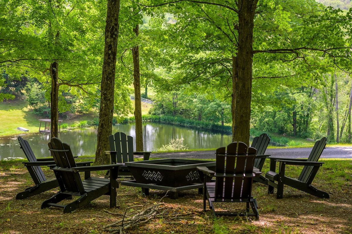 A circular fire pit surrounded by dark Adirondack chairs, nestled under a canopy of green trees with a glimpse of a pond in the background, perfect for outdoor gatherings.